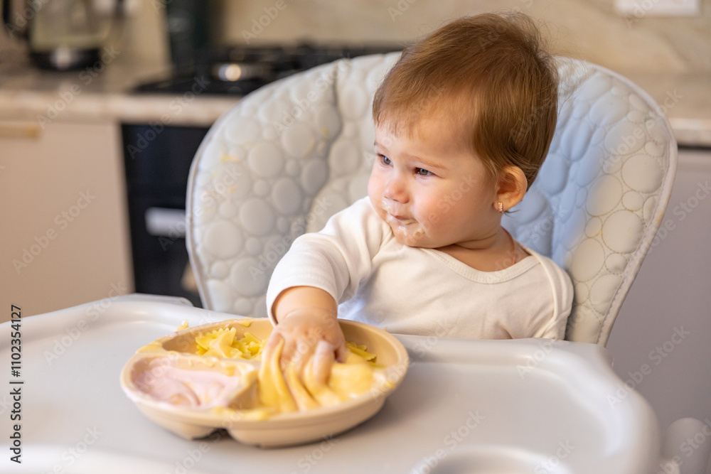 Cute little toddler kid girl tasting and enjoying the greek yogurt and pasta sitting in baby chair with messy face. Self-feeding concept. First solid food. Children meal concept. High quality photo Cute little toddler kid girl tasting and enjoying the greek yogurt and pasta sitting in baby chair with messy face. Self-feeding concept. First solid food. Children meal concept. High quality photo