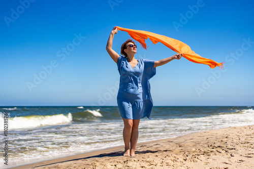 Summer vacation. Beautiful mature woman holding scarf fluttering in wind standing on sandy beach on sunny day. Front view