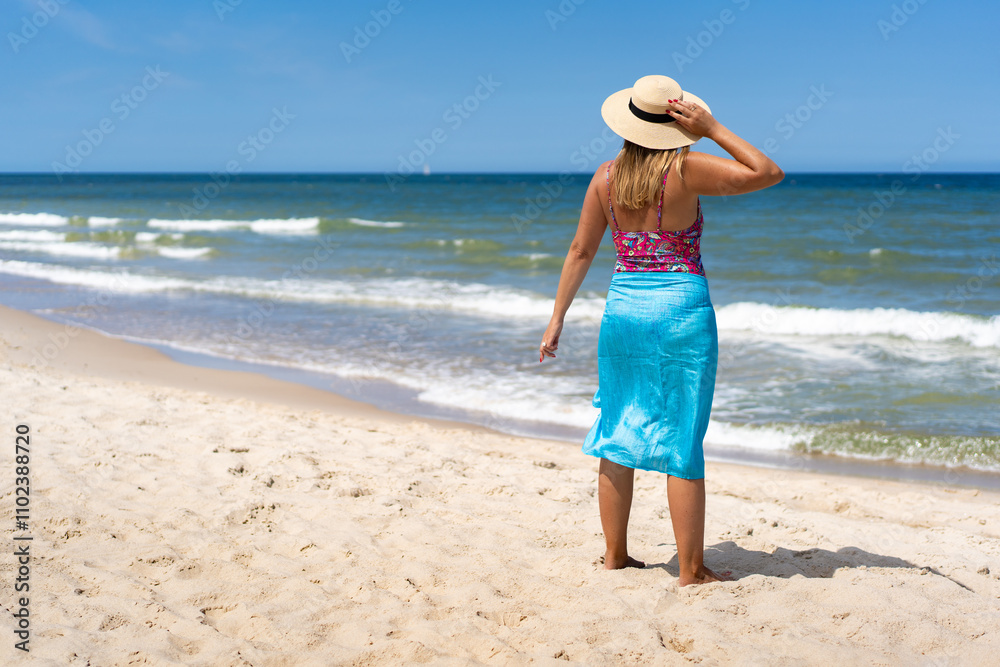 Obraz premium Summer vacation. Tanned mature woman in swimsuit, pareo and sun hat standing by shore on sandy beach. Back view