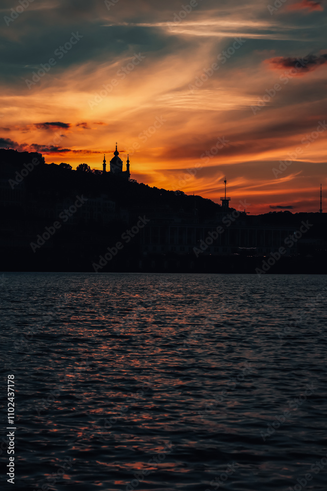 Naklejka premium Contours city buildings and silhouette of a church at sunset with vibrant orange and purple hues over a calm river. 