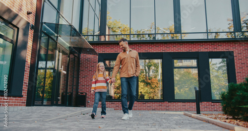 Cute Caucasian girl with backpack and glasses holding hand of her loving father. Walking together back home from school. Trees visible mirroring in tall windows of modern building.