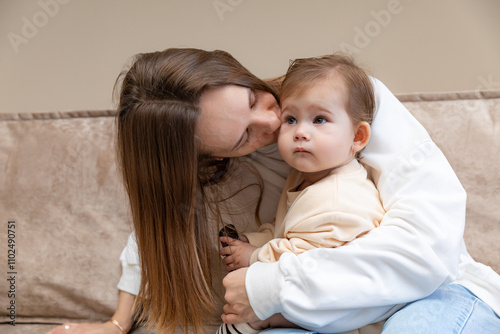 Young caring mother hugged her little daughter, pressing her to her chest. Woman holding a newborn baby in her arms on a sofa at home. Smiling mother hugging a small newborn baby showing love