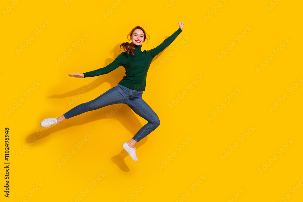 Full length photo of adorable lovely girl wear green pullover jumping high risig fist empty space isolated yellow color background Full length photo of adorable lovely girl wear green pullover jumping high risig fist empty space isolated yellow color background