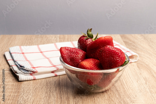 bol de vidrio con frutillas rojas maduras sobre una mesa de madera, junto a un paño de cocina photo