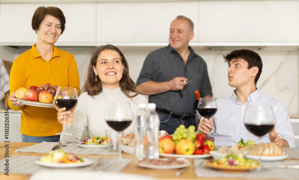 Cheerful young girl sitting at table with husband and senior parents during cozy family celebration over kitchen table served with wine and light snacks Cheerful young girl sitting at table with husband and senior parents during cozy family celebration over kitchen table served with wine and light snacks