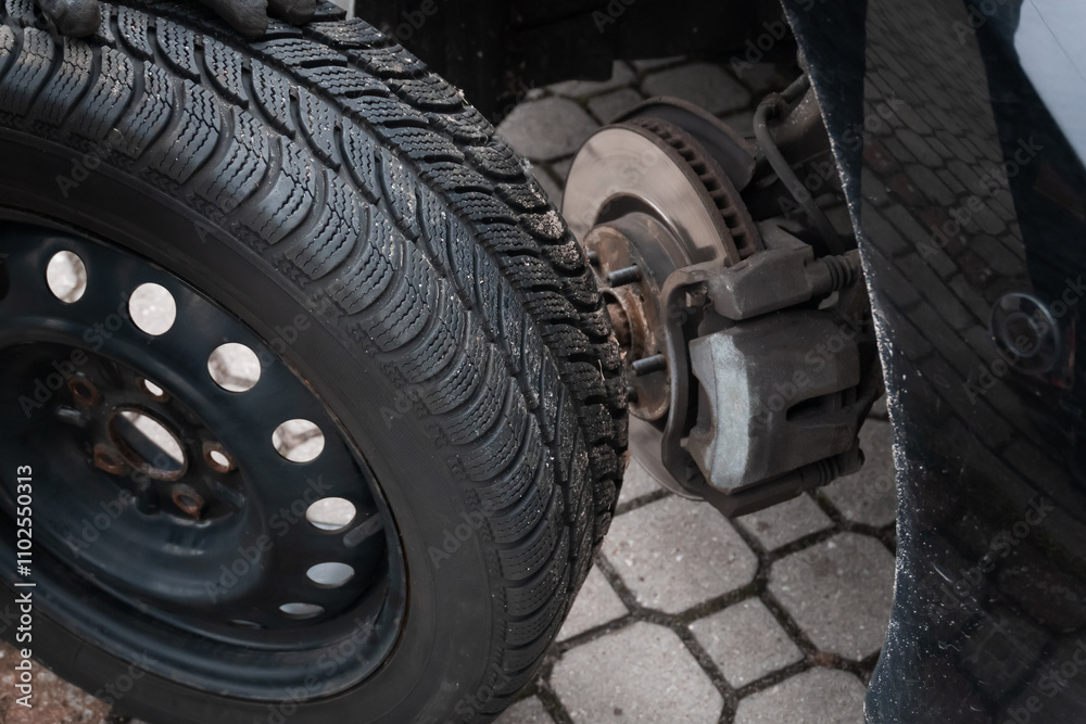 A car tire replacement in progress. A person wearing black gloves is handling the removed wheel near the vehicle, revealing the exposed brake disc and suspension system. A car tire replacement in progress. A person wearing black gloves is handling the removed wheel near the vehicle, revealing the exposed brake disc and suspension system.