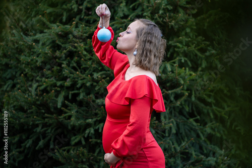 It is a boy. Pregnant woman is expecting baby boy. Future mother holds blue ball bauble. Winter pregnancy. Woman in red dress stands near evergreen fir tree