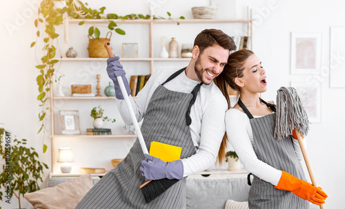 Great house cleaning together and rock band. Cheerful husband plays on broom, wife in apron sings in imaginary microphone in mop, in interior of modern living room with cleaning supplies, free space photo