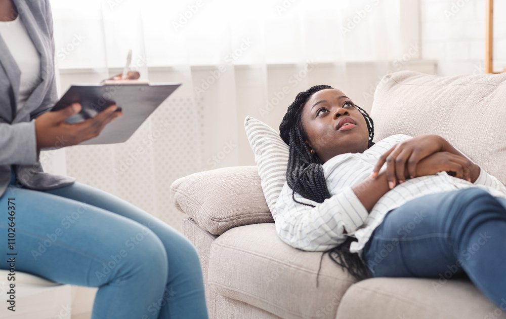 At the psychologist. Depressed young afro woman lying on couch during psychotherapy session while doctor taking notes, free space At the psychologist. Depressed young afro woman lying on couch during psychotherapy session while doctor taking notes, free space