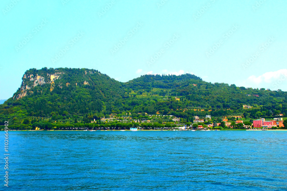 Majestic view at Bardolino on the eastern shore of Lake Garda with Rocca di Garda, a mountainous rocky cliff filled with vegetation, different lightly-colored buildings, Hotel Sportsman and waters Majestic view at Bardolino on the eastern shore of Lake Garda with Rocca di Garda, a mountainous rocky cliff filled with vegetation, different lightly-colored buildings, Hotel Sportsman and waters
