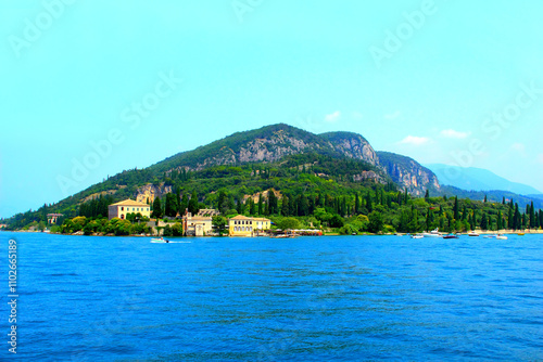 Perfect scenery with Punta San Vigilio on Lake Garda, vegetation and cypress trees extending on the shore near a multitude of motorboats, reaching pastel-colored buildings under Monte Luppia