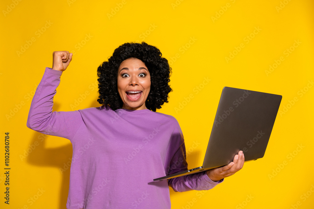 Portrait of pretty young woman hold netbook raise fist wear violet pullover isolated on yellow color background Portrait of pretty young woman hold netbook raise fist wear violet pullover isolated on yellow color background