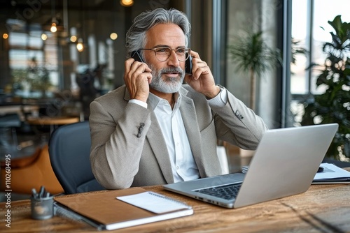 Middle aged businessman on phone in modern office working on laptop at desk communicating with client