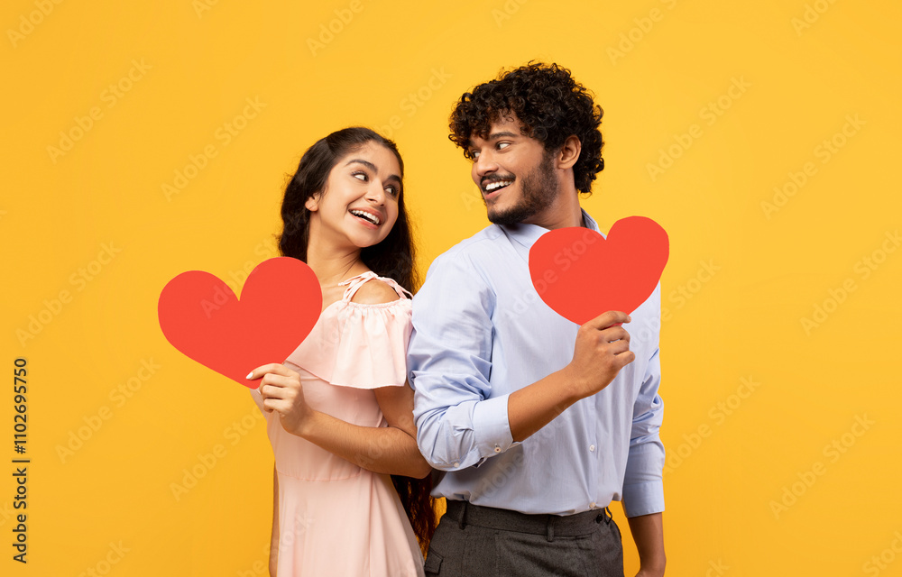 Valentine's holiday. Portrait of indian lovers holding red paper hearts and looking at each other, standing on yellow studio background. Romantic relationship concept Valentine's holiday. Portrait of indian lovers holding red paper hearts and looking at each other, standing on yellow studio background. Romantic relationship concept