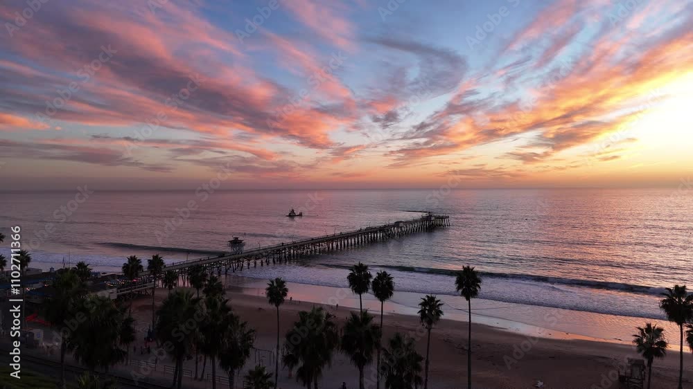 A drone-captured aerial view of the San Clemente Pier during a colorful sunset, showcasing vibrant orange and pink clouds reflecting over the Pacific Ocean. Palm trees line the beach below