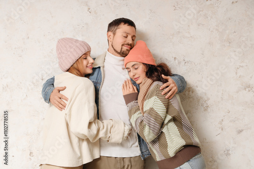 Man and two beautiful women in autumn clothes hugging near white wall. Polyamory concept photo