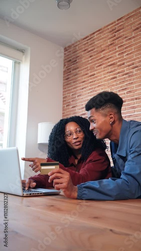 Vertical. Smiling African American couple using a laptop and credit card to make an online purchase, enjoying convenient shopping at home in a cozy modern setting with a brick wall background.