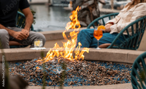 A fire in a fire pit with a man and a woman sitting with cocktails diffused in the background near water