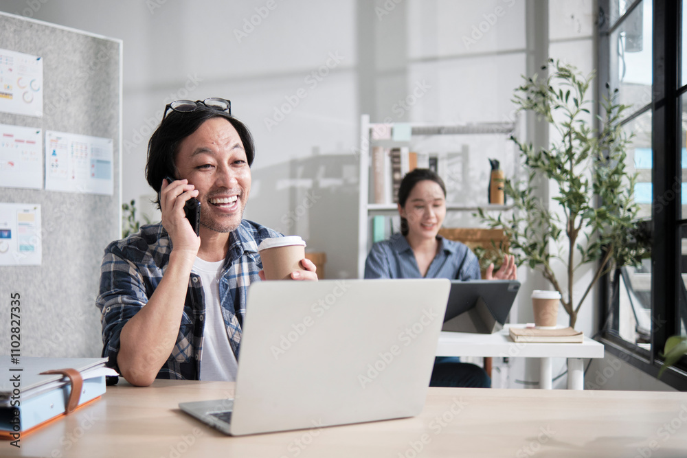 Asian startup partners working in a casual small business office, male worker talking on mobile phone with customers, online service jobs, female colleague behind, and E-commerce SME coworker team. Asian startup partners working in a casual small business office, male worker talking on mobile phone with customers, online service jobs, female colleague behind, and E-commerce SME coworker team.