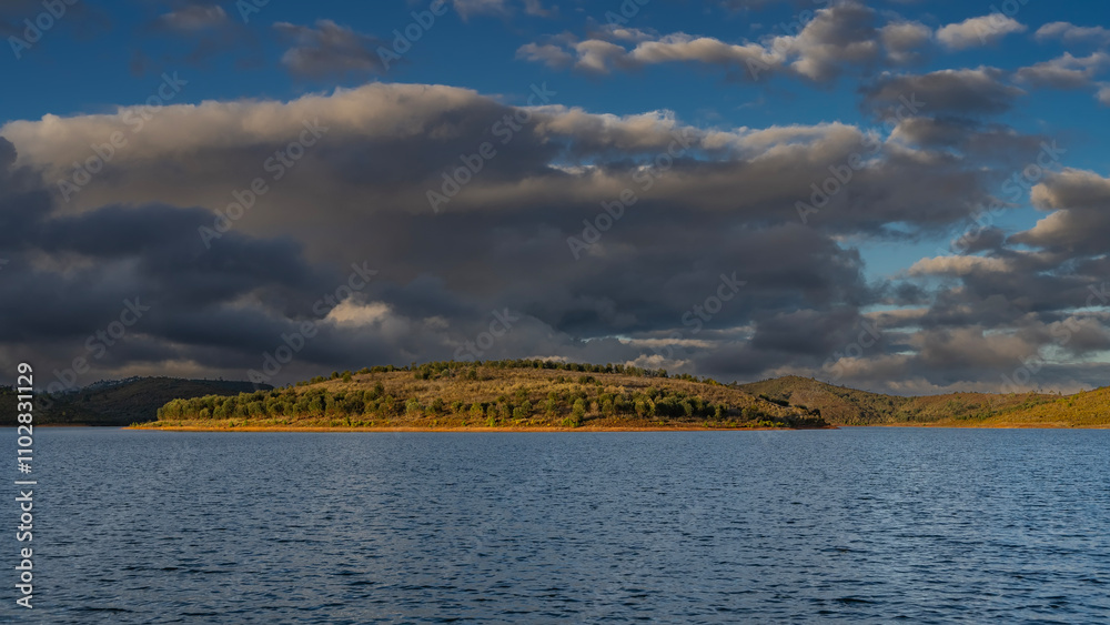 Green trees and yellowed grass grow on the hilly shores of the lake. Ripples on the blue water. The clouds in the sky are highlighted. The evening golden hour. Madagascar. Mantasoa Lake. Green trees and yellowed grass grow on the hilly shores of the lake. Ripples on the blue water. The clouds in the sky are highlighted. The evening golden hour. Madagascar. Mantasoa Lake.