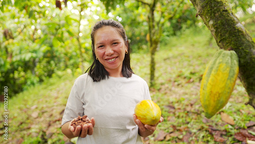 Portrait of smiling southeast asian woman farmer in cacao farm holding harvested pod in one hand and processed beans in the other