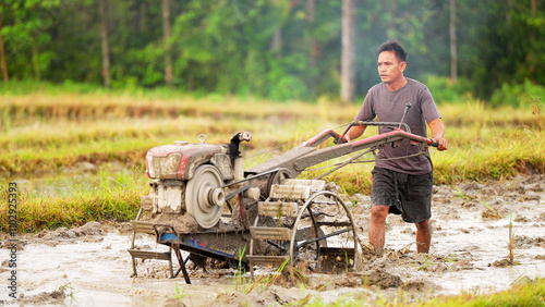 Southeast asian male rice farmer operating a hand tractor in a muddy field on terraced farmland, smallholder farming practices