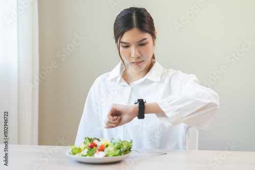 Intermittent fasting, diet healthy asian young woman dietary watching wrist watch, temptation hungry of brunch food in morning on table at home, waiting time to eat salad but not yet time first meal.