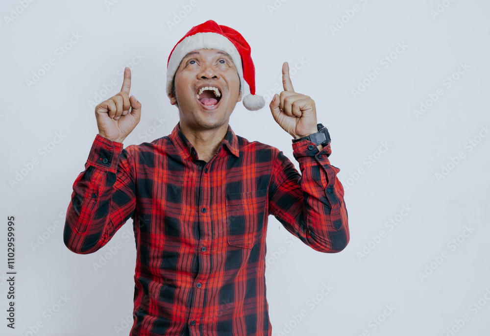 Merry young asian man wearing red checkered shirt and  santa hat posing point index finger above indicate on workspace area isolated on white background. Happy New Year, celebration holiday concept.