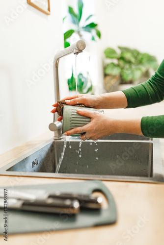 Cleaning, woman in green dress with green ceramic cup in kitchen for hygiene, routine and coffee mug. Female person, washing with water for disinfecting, protection and sink at home 