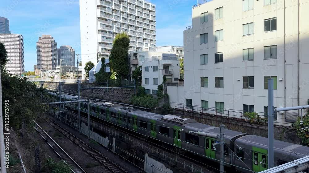 A green commuter train moves through a modern urban area with tall buildings in Tokyo, Japan A green commuter train moves through a modern urban area with tall buildings in Tokyo, Japan
