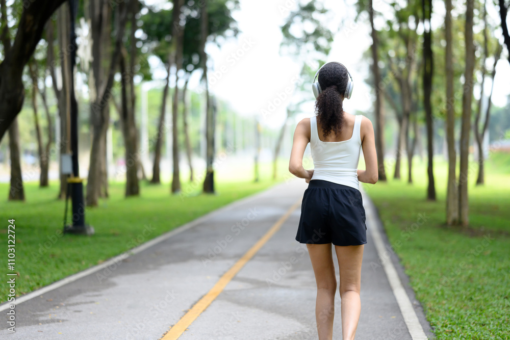 Back view of young sportswoman with headphones jogging through a public park Back view of young sportswoman with headphones jogging through a public park
