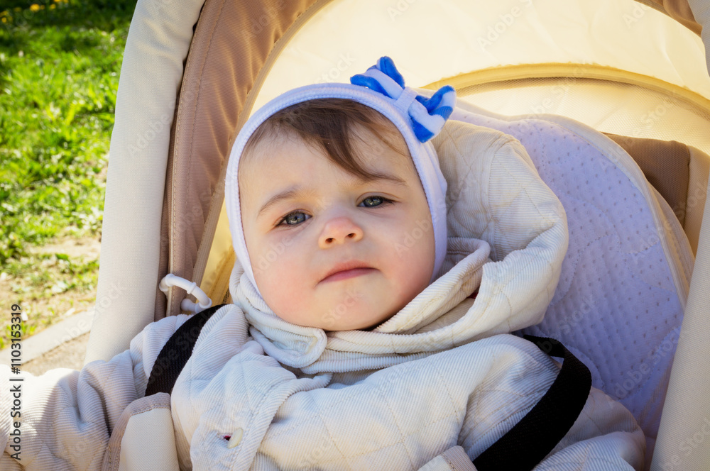 A small child or baby or girl or toddler in a jacket and hat sits in a baby carriage on a walk. Close-up portrait of a child A small child or baby or girl or toddler in a jacket and hat sits in a baby carriage on a walk. Close-up portrait of a child