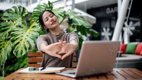 A relaxed, carefree Asian woman stretches her arms while sitting in front of her laptop outdoors.