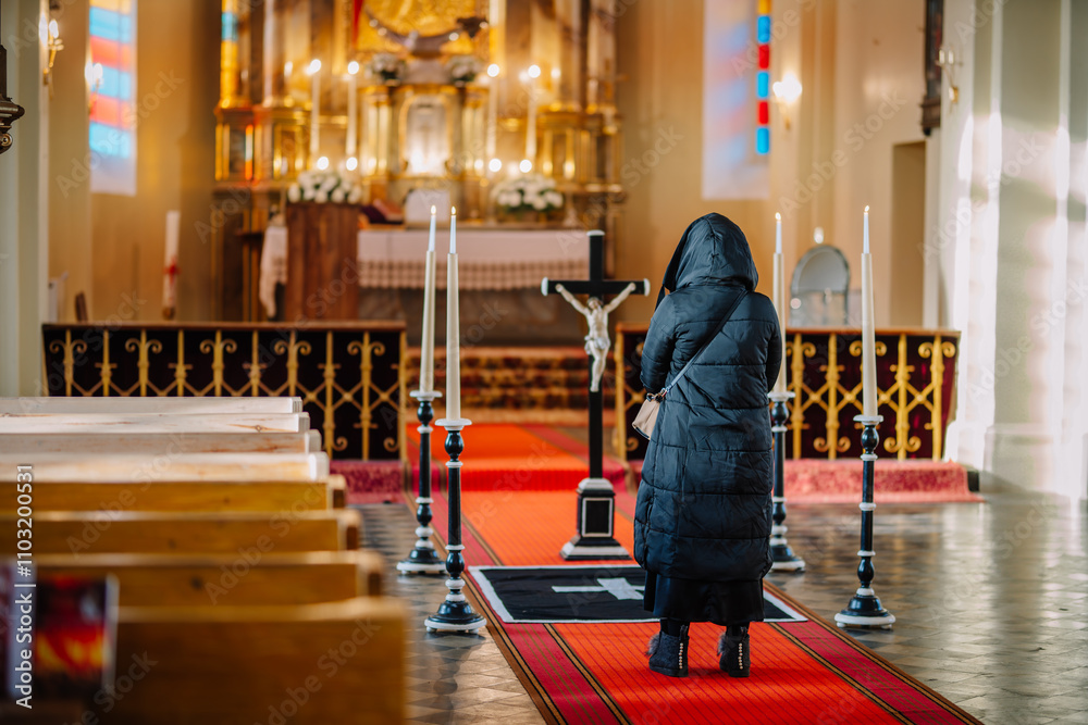 Obraz premium A woman in a black coat stands in a church during a funeral, facing the altar with lit candles and a crucifix, reflecting somber emotions..