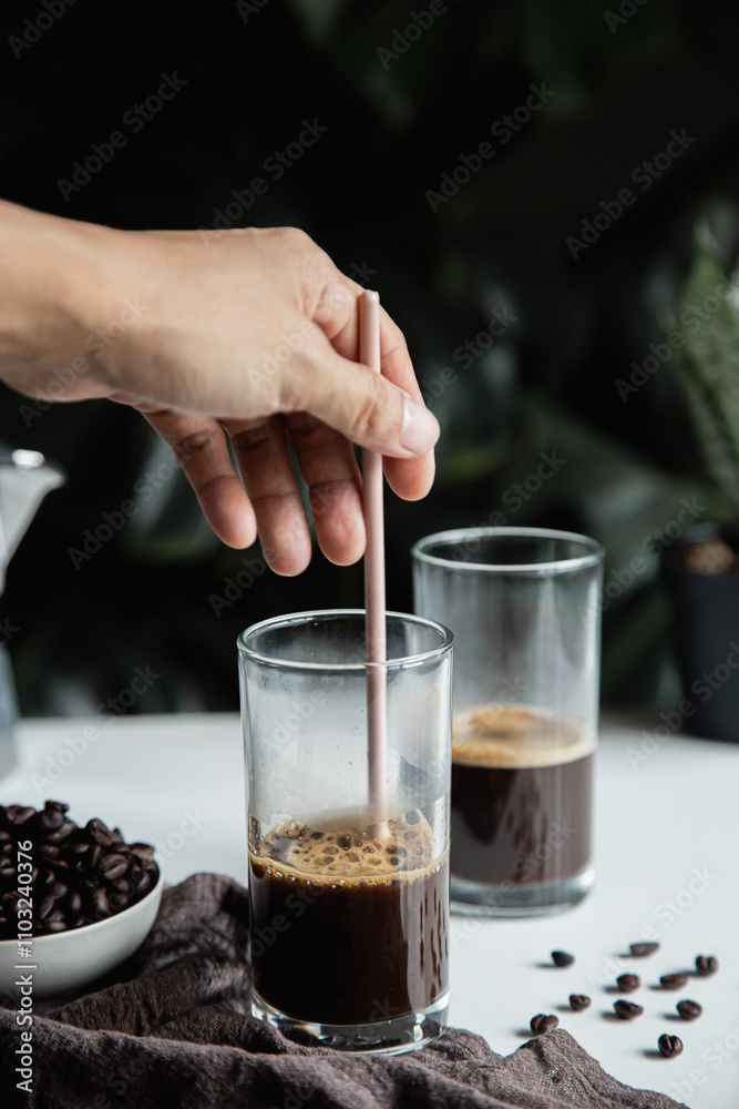 person's hand is using a spoon to mix instant coffee on a glass to prepare an iced latte. vertical photo person's hand is using a spoon to mix instant coffee on a glass to prepare an iced latte. vertical photo