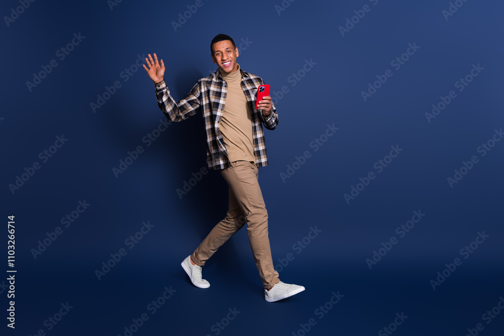 Full length photo of funky cool guy dressed plaid shirt reading modern device waving arm hi emtpy space isolated blue color background Full length photo of funky cool guy dressed plaid shirt reading modern device waving arm hi emtpy space isolated blue color background