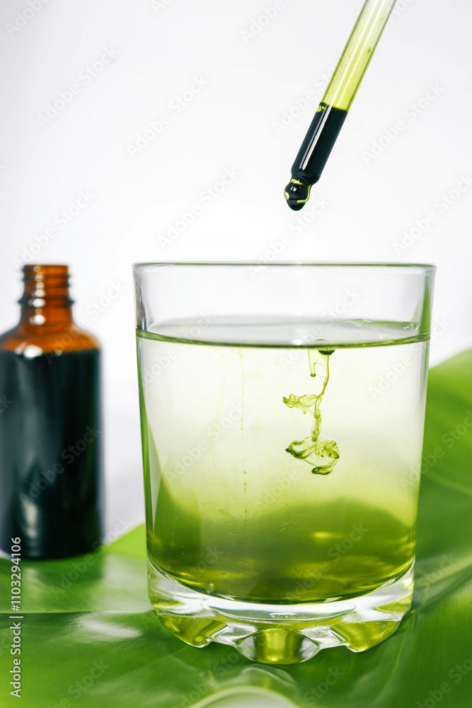 Dropper releasing green liquid into a glass of water with a leaf underneath and a brown glass bottle in the background. Represents natural ingredients, organic products Dropper releasing green liquid into a glass of water with a leaf underneath and a brown glass bottle in the background. Represents natural ingredients, organic products