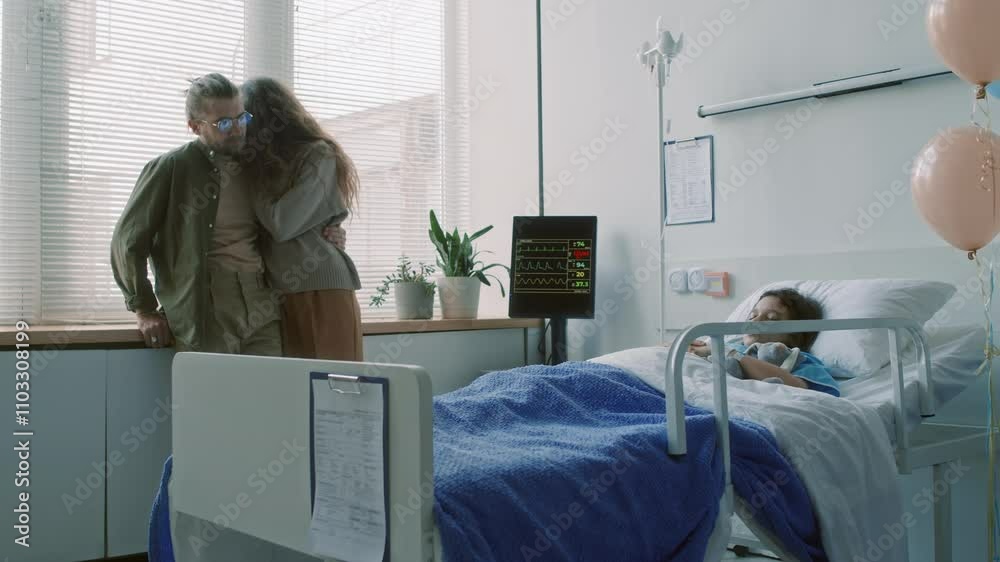 Full shot of worried woman hugging husband while staying with sick daughter lying in hospital bed in coma in ward decorated with balloons Full shot of worried woman hugging husband while staying with sick daughter lying in hospital bed in coma in ward decorated with balloons