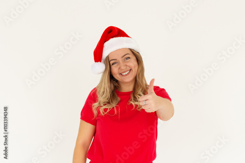 Young funny european woman in red t-shirt and christmas hat showing thumb up and smiling. Happy New Year and Merry Christmas concept