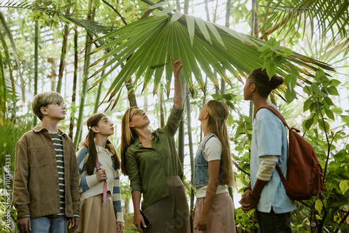 Young female tour guide working in botanical garden greenhouse showing sabal palm leaves to group of teenagers photo