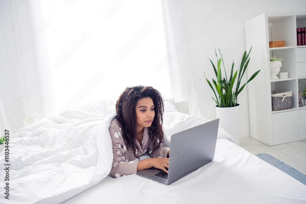 Young woman in pajamas using laptop on bed in bright morning light, enjoying comfort of home Young woman in pajamas using laptop on bed in bright morning light, enjoying comfort of home