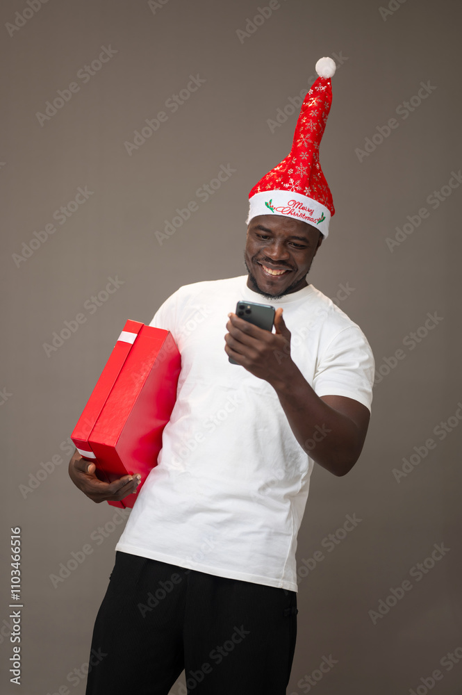 Young African American Man Wearing a Christmas Hat, Smiling, Holding a Christmas Gift Box, and Excitedly Going Through His Smartphone Young African American Man Wearing a Christmas Hat, Smiling, Holding a Christmas Gift Box, and Excitedly Going Through His Smartphone