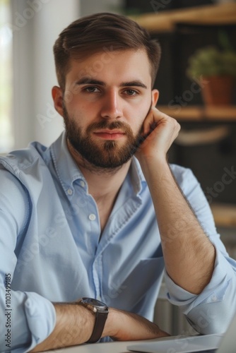 A bearded man in a blue shirt, resting his chin on his hand and looking away.