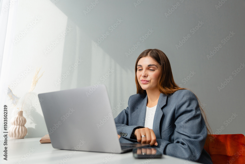 Woman in a gray blazer is looking at her laptop and smiling during an online conference. The workplace is well lit and a vase of dried plants adds aesthetic appeal. Copy space. Woman in a gray blazer is looking at her laptop and smiling during an online conference. The workplace is well lit and a vase of dried plants adds aesthetic appeal. Copy space.