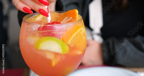 Fresh summer aperol spritz, mimosa cocktail with ice and fruits, oranges, apples and strawberries. Hands of a young girl with a beautiful red manicure slowly stir a drink with a straw. Close up shot