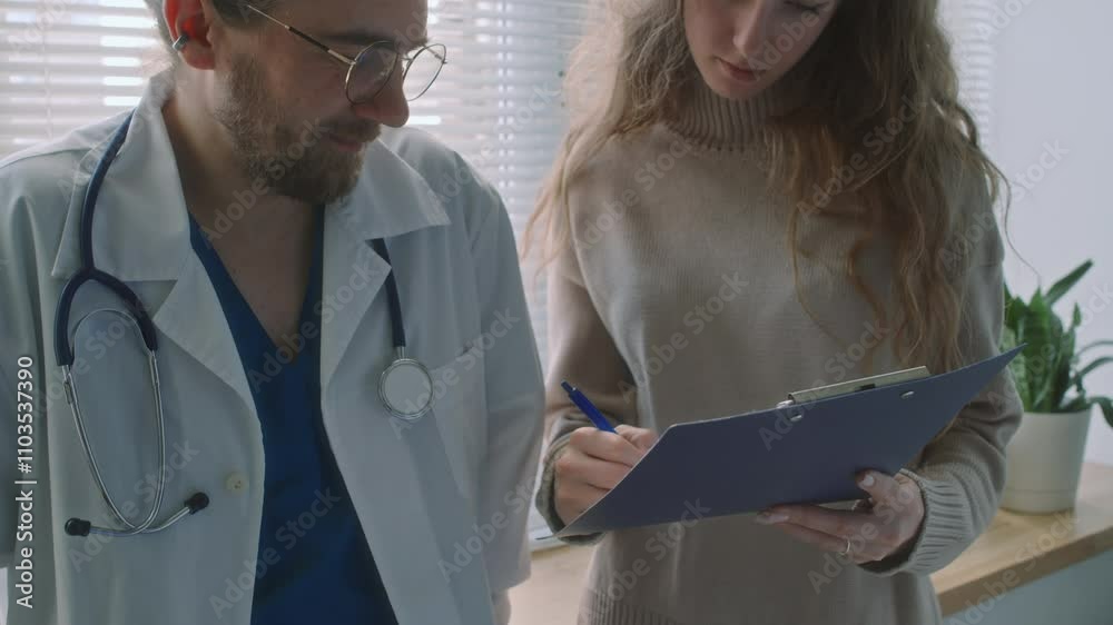 Tilt shot of curly-haired Caucasian woman discussing treatment plan with male doctor and signing it in modern clinic