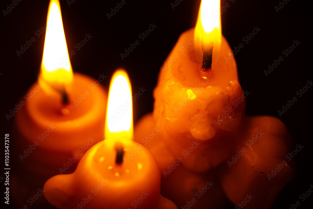 Three lit candles light up the dark background with soft, warm flames. The wax appears slightly melted, creating a calming and serene atmosphere. The image features close-up details. Three lit candles light up the dark background with soft, warm flames. The wax appears slightly melted, creating a calming and serene atmosphere. The image features close-up details.