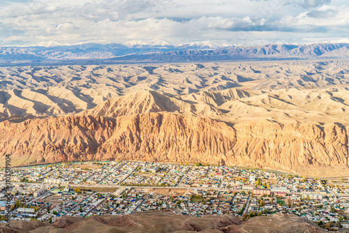 A striking aerial view of a small town nestled at the foot of rugged red cliffs and surrounded by an expansive desert-like landscape. The intricate patterns of the rolling hills and distant mountains 