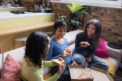 Female friends sharing takeout pizza sitting on the couch in their loft apartment photo