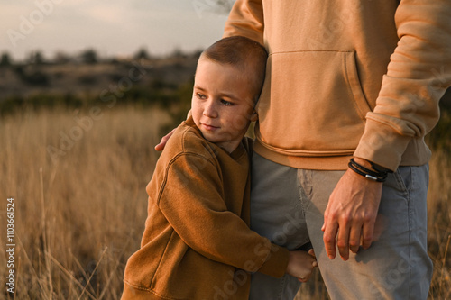 A boy gazes at the sunset while lovingly holding his father’s leg. The warm glow of the setting sun creates a tender, emotional moment between father and child. photo